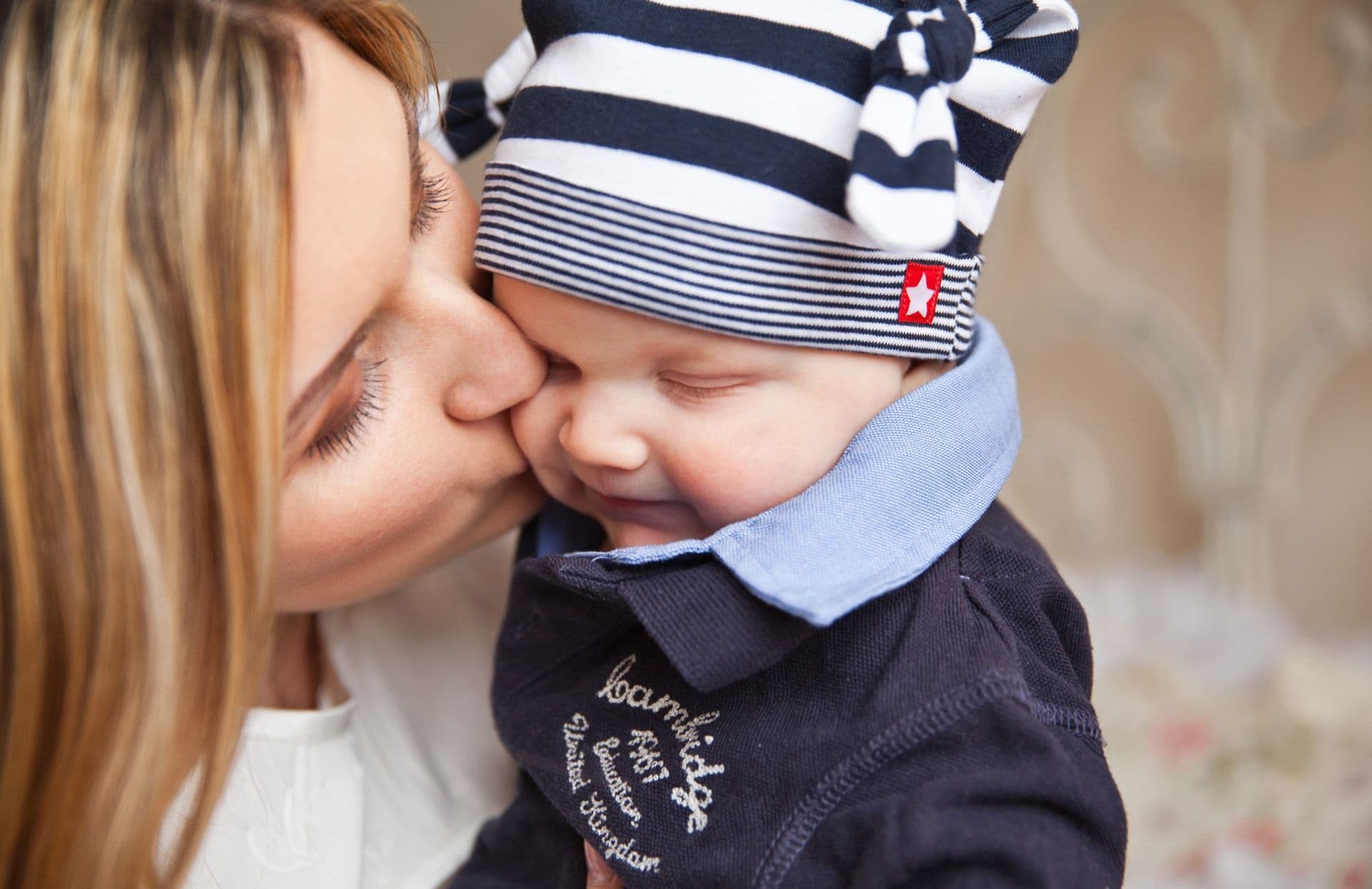 Mãe beijando a bochecha de um bebê de gorro, que sorri de olhos fechados